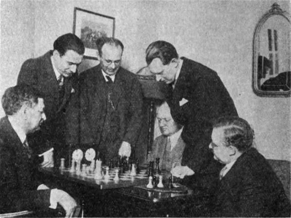 Julius Finn (links), Vorsitzender des Komitees, spielt mit Rudolf Spielmann. Zuschauer, von links nach rechts, sind Dr. N. L. Lederer, A. Nimzowitsch, Dr. A. Aljechin und Dr. M. Vidmar.